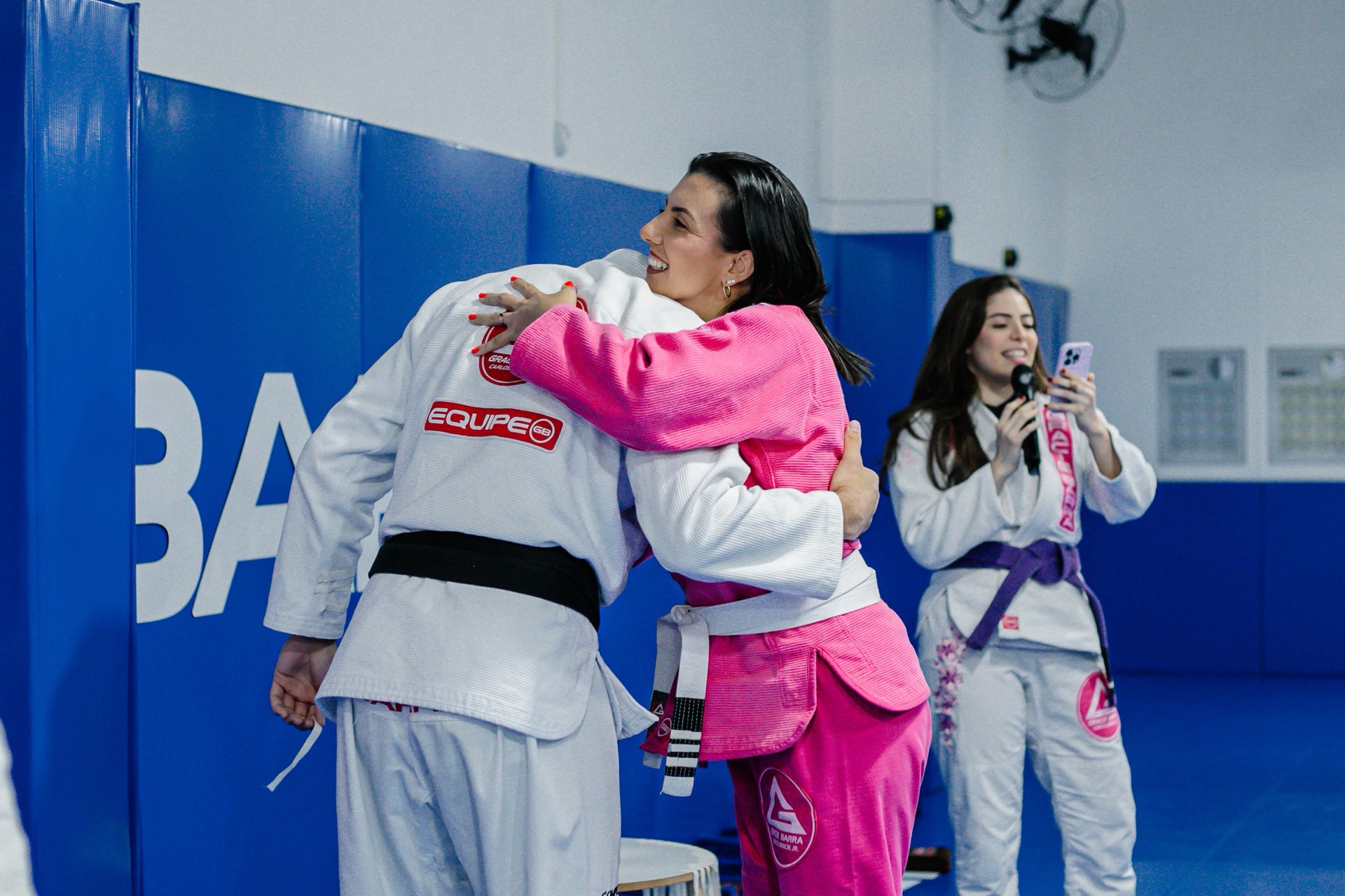 Instrutor faixa preta abraçando uma aluna de kimono rosa no tatame