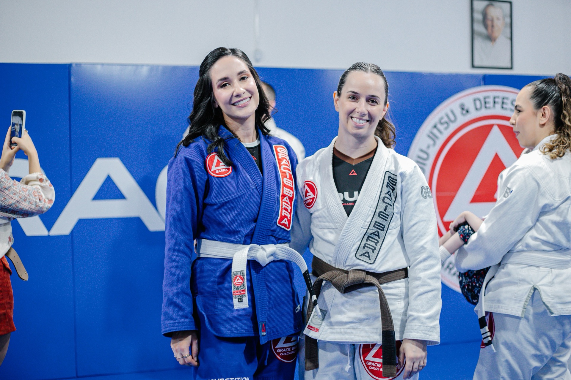 Duas alunas de faixas coloridas sorrindo diante do escudo da escola