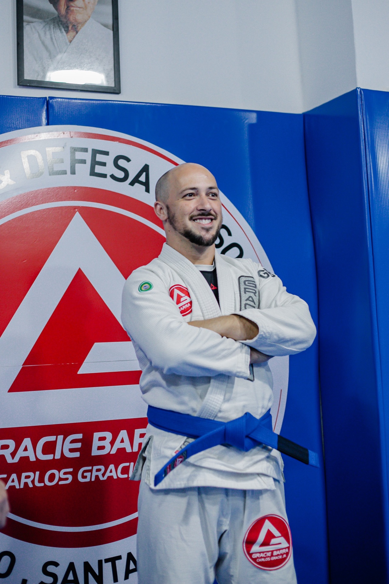 Aluno de faixa azul com braços cruzados diante do escudo Gracie Barra Tubarão pintado na parede