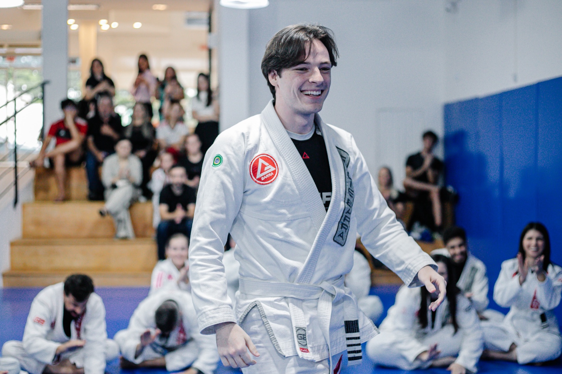 Aluno de faixa branca sorrindo em pé no tatame durante a aula