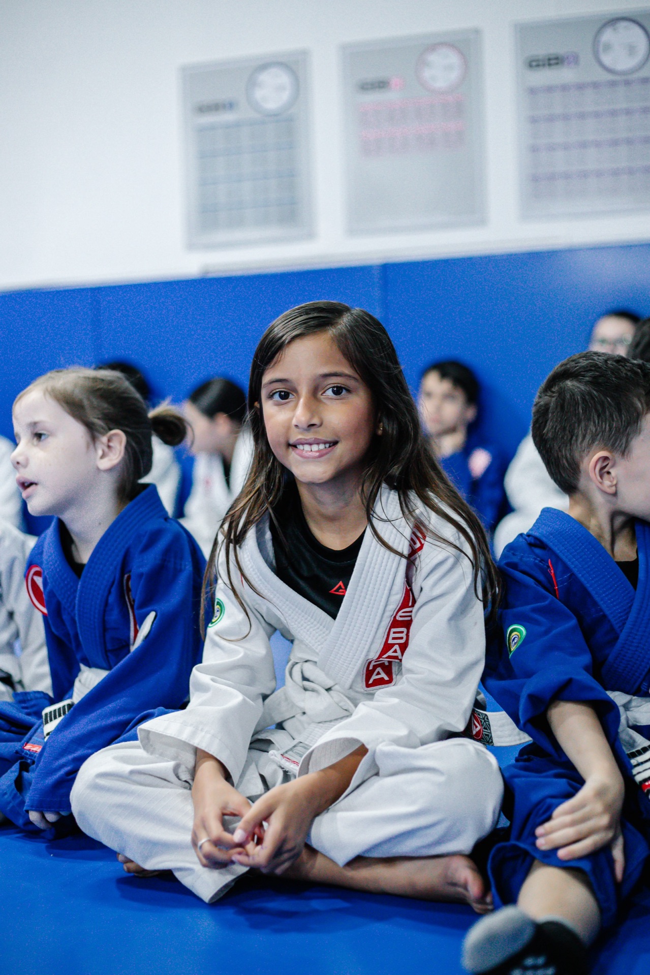 Menina de kimono branco sentada no tatame, sorrindo, com outras crianças da turma Kids ao lado