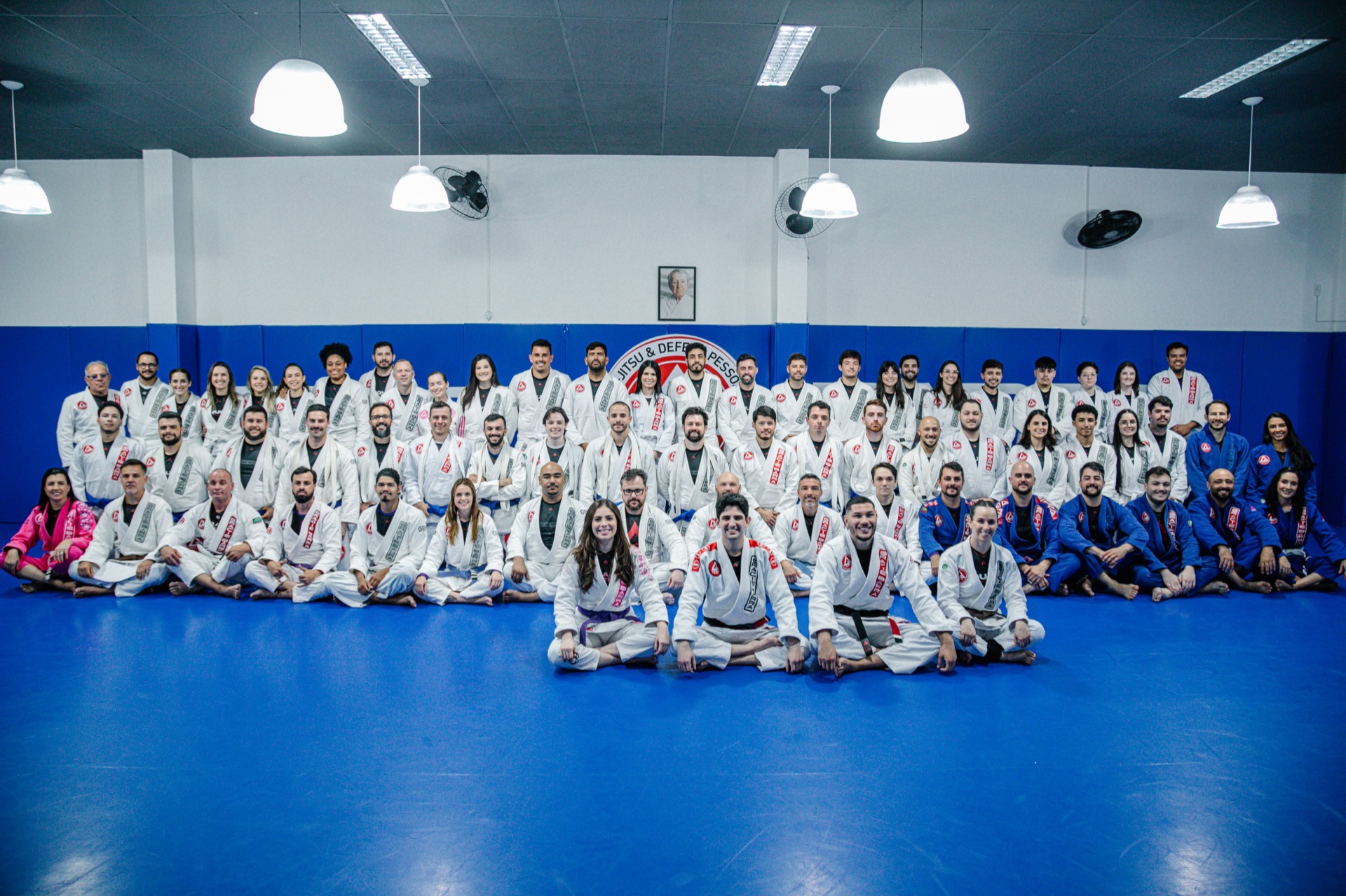 Alunos e instrutores da Gracie Barra Tubarão reunidos no tatame azul, de kimono, crianças, mulheres e homens de todas as faixas